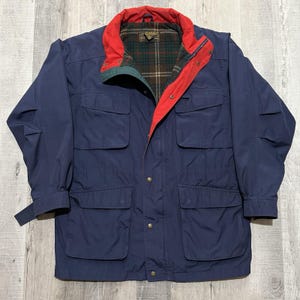 May include: A navy blue jacket with a red collar and green accents. The jacket has four front pockets and a plaid lining. The sleeves have adjustable cuffs. The jacket is buttoned up and appears to be made of a durable material.