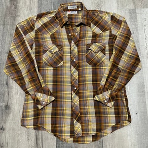 May include: A brown and yellow plaid long-sleeved shirt with a button-down collar and two chest pockets. The shirt has a label that reads "Enjoy" in white letters on a red background.