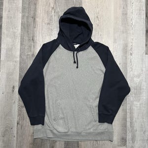 May include: A gray hooded sweatshirt with navy blue raglan sleeves. The sweatshirt has a front pocket and a drawstring at the hood. The garment is made of a soft, comfortable material.