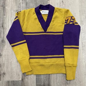 May include: A vintage gold and purple sweater with a V-neck design. The sweater features purple sleeves and horizontal stripes, with the number "77" on the right sleeve. The label reads "Barry".