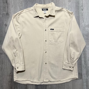 May include: A light beige button-down shirt with long sleeves. The shirt has a collared neckline, a chest pocket, and the brand name "GUESS" is visible on the collar and pocket. The shirt is made of a sturdy fabric.