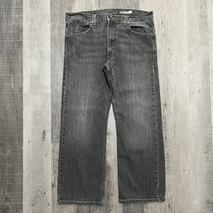 May include: A pair of faded black denim jeans with a classic five-pocket design. The jeans have a straight leg cut and show subtle wear marks. The metal rivets and button closure are visible. The jeans are displayed on a light gray wooden surface.