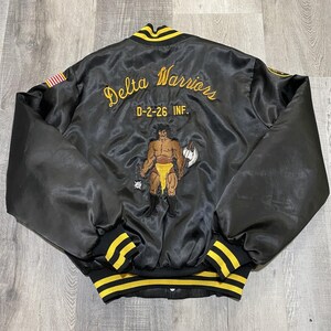 May include: A black satin bomber jacket with yellow striped ribbed trim at the collar, cuffs, and hem. The back features yellow embroidered text reading Delta Warriors and D-2-26 INF, with a central graphic of a warrior figure holding an axe.