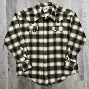 May include: A long-sleeved, button-down shirt in a black and cream plaid pattern. The shirt features a pointed collar, two chest pockets with decorative stitching, and pearl snap closures. The cuffs also have pearl snap closures.