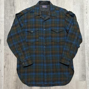 May include: A long-sleeved, button-down shirt in a plaid pattern of blue, brown, and green. The shirt has two chest pockets and a quilted collar. The fabric appears to be a heavy wool or wool blend. The shirt is laid flat on a light gray surface.