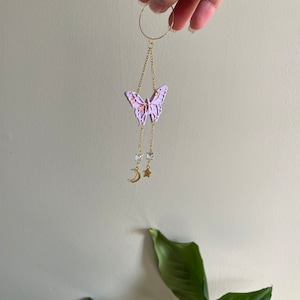 May include: A gold chain with a purple butterfly charm, a crescent moon, and a star charm. The chain is attached to a gold hoop.