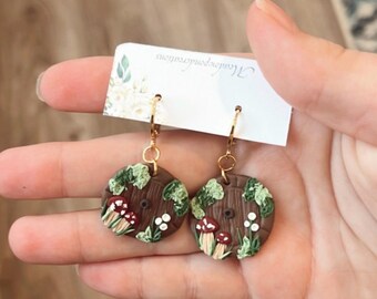 May include: Handmade polymer clay earrings with a forest theme. The circular earrings have a brown base, green leaves, red and white mushrooms, and small white flowers. Gold-coloured ear wires are attached. The earrings are held by a hand.