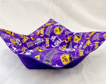 May include: A purple and yellow fabric bowl with a square base and flared sides. The interior fabric features a repeating pattern of LSU Tigers logos and helmets. The exterior is a solid purple color.