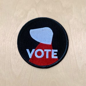 Puede incluir: Un parche bordado negro y rojo con una silueta blanca de la cabeza de una persona y la palabra "VOTE" en blanco.