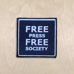 May include: A black square patch with white text that reads "FREE PRESS FREE SOCIETY".
