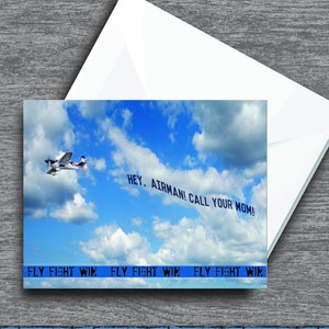 May include: A blue and white airplane flies through a cloudy sky with a banner that reads "HEY, AIRMAN! CALL YOUR MOM!" The banner is attached to the airplane with a rope. The words "FLY FIGHT WIN" are repeated three times in a blue banner at the bottom of the image.