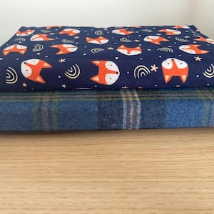 Puede incluir: Dos piezas de tela dobladas. La pieza superior es azul marino con un patrón repetido de zorros naranjas, detalles blancos y estrellas. La pieza inferior es un estampado de cuadros azules con rayas verdes y marrones. Las telas están apiladas sobre una superficie de madera.