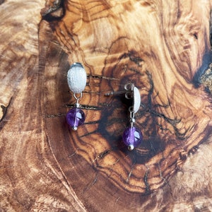 May include: A pair of silver earrings with a textured oval charm and a dangling purple amethyst bead.