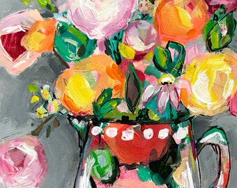 May include: A colorful bouquet of pink, yellow, and orange flowers in a white pitcher with red and white polka dots. The pitcher has a handle and is sitting on a pink and gray surface.