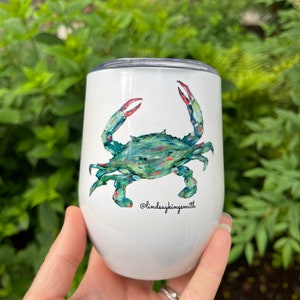 May include: White stainless steel tumbler with a green and blue crab illustration. The crab has red claws and is painted in a watercolor style. The tumbler has a black text that reads "@lindsayskingsmith".