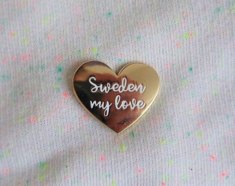 Könnte beinhalten: Goldene herzförmige Anstecknadel mit dem Text "Sweden my love" in weiß.