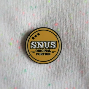 Può includere: Uno spillo in smalto dorato e nero con il testo "SNUS ORIGINAL PORTION" e tre stelle.