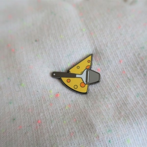 May include: A yellow enamel pin shaped like a slice of cheese with a small silver cheese grater on top.