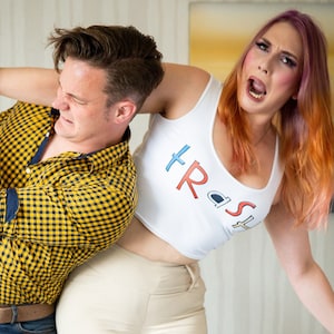 May include: A man and a woman are playfully wrestling. The woman is wearing a white vest top with the word "Trash" printed on it in red, blue, and white letters. The man is wearing a yellow and black checked shirt and blue jeans.