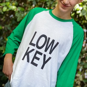 May include: White and green raglan-style long-sleeve shirt with the text "LOW KEY" printed in black. The shirt has green sleeves and a white body. The person is smiling in front of a green leafy background.
