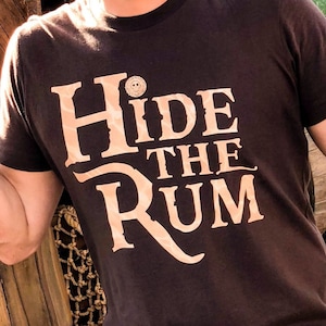May include: A man wearing a brown t-shirt with the text "Hide the Rum" in a gold font. He is standing in front of a pile of wooden barrels.