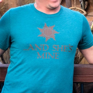 Puede incluir: Un hombre lleva una camiseta azul turquesa con un gráfico de sol dorado y el texto "...and she's mine". Está señalando una pantalla de pescado seco colgado de una estructura de madera.