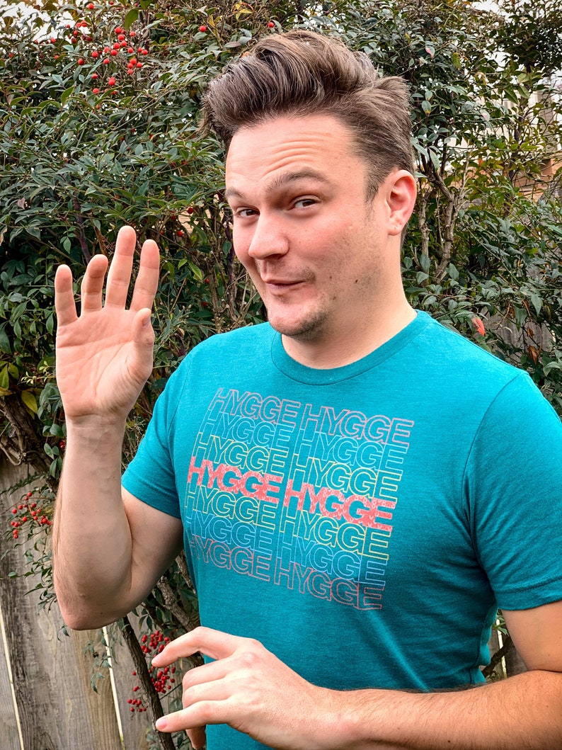 May include: A man wearing a teal blue t-shirt with the word "hygge" repeated in a rainbow pattern. He is smiling and waving with his right hand.