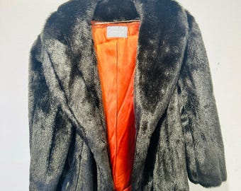 May include: A black faux fur coat with a wide collar and an orange lining. The coat is hanging on a copper-coloured hanger against a white wall. The coat has a classic design.