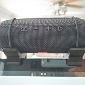 May include: A black portable Bluetooth speaker with a fabric mesh covering and a rubber strap. The speaker has buttons for play, pause, volume up, volume down, and repeat. The speaker is mounted on a black stand.