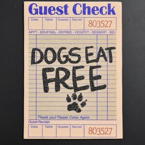 May include: A rectangular tan guest check pad with blue grid lines and text. The top reads Guest Check 803527. The center features the black text DOGS EAT FREE above a black paw print. The bottom includes the text Thank you! Please Come Again.