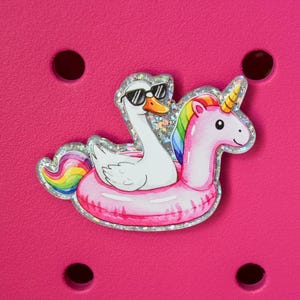 May include: A whimsical charm featuring a white swan wearing sunglasses and a pink unicorn, both riding a pink inflatable ring. The charm has a glittery silver border and is set against a bright pink background.