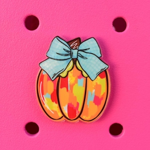 May include: A colorful pumpkin charm with a light blue bow. The pumpkin is orange with a mix of yellow, red, and blue brushstrokes. The charm is set against a bright pink background with four dark brown circular holes.