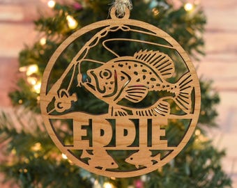 May include: Wooden Christmas ornament with a fishing theme. The round ornament features a fish, fishing rod, and the name "EDDIE" cut out. It is hanging on a Christmas tree. The ornament is made of light brown wood and has a natural wood grain.