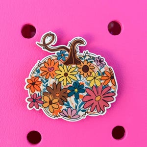 May include: A decorative pumpkin-shaped charm with a floral design. The charm features a brown stem and is filled with colorful flowers in shades of pink, orange, yellow, and blue. The charm is set against a bright pink background.