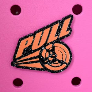 May include: A pink charm with the word "PULL" in orange, outlined with black glitter. The charm also features an orange graphic of a wheel with black glitter accents. The charm is set against a pink background with four dark red holes.