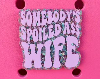 Könnte beinhalten: Ein hellvioletter Schuhanhänger mit dem Text "SOMEBODY'S SPOILED ASS WIFE" in einer Blasen-Schriftart. Der Anhänger hat einen glitzernden, irisierenden Hintergrund und ist vor einem hellrosa Hintergrund mit vier runden Löchern platziert.