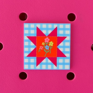 May include: A square, decorative patch featuring a star design with a floral center. The star is hot pink, set against a light blue and white gingham pattern. The center is orange with colorful flowers.