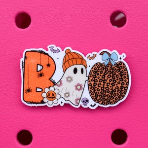 May include: A Halloween-themed charm with the word "BOO" spelled out with an orange letter "B" decorated with spiders and a web, a ghost wearing an orange beanie, and a leopard-print pumpkin with a blue bow.
