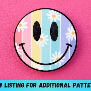 May include: A round, colorful smiley face design with a rainbow striped background and daisy accents. The design has a black outline and is set against a pink background. Text at the bottom reads "VIEW LISTING FOR ADDITIONAL PATTERNS".