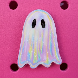 May include: A holographic iridescent ghost charm with black eyes. The ghost is a light purple color with a white outline. The charm is on a pink background.