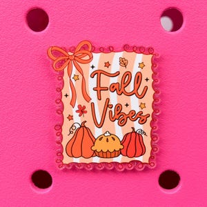 May include: A decorative charm with the words "Fall Vibes" in orange script, surrounded by pumpkins, a pie, and fall-themed accents. The charm has a scalloped border and a bow at the top, set against a bright pink background.