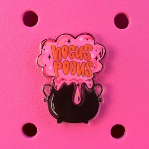 May include: A black cauldron charm with the words "Hocus Pocus" in orange letters, surrounded by a pink cloud with stars. The charm is set against a bright pink background.