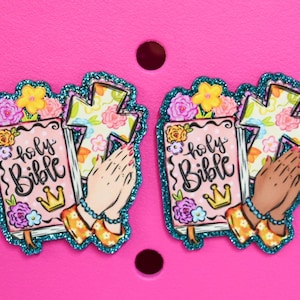 May include: Two decorative charms featuring a pink Bible with the words "holy Bible" and a gold crown. The charms also include a cross, praying hands, and floral accents, outlined with teal glitter. Set against a pink background.