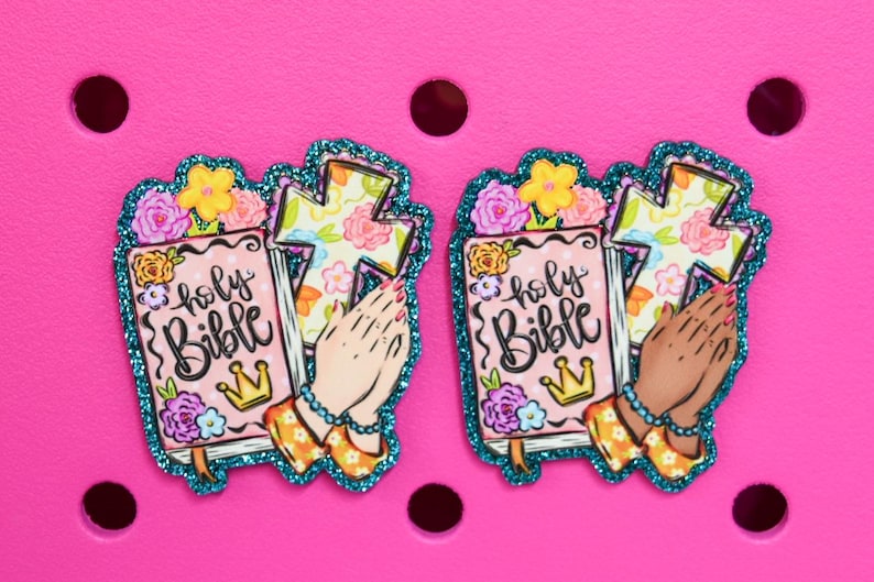 May include: Two decorative charms featuring a pink Bible with the words "holy Bible" and a gold crown. The charms also include a cross, praying hands, and floral accents, outlined with teal glitter. Set against a pink background.