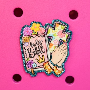 May include: A decorative charm featuring a pink Bible with the words "holy Bible" and a gold crown, a floral cross, and praying hands. The charm has a teal glitter border and is set against a bright pink background.