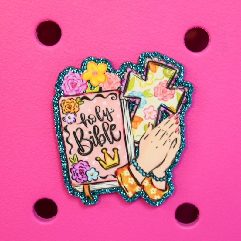 May include: A decorative charm featuring a pink Bible with the words "holy Bible" and a gold crown, a floral cross, and praying hands. The charm has a teal glitter border and is set against a bright pink background.