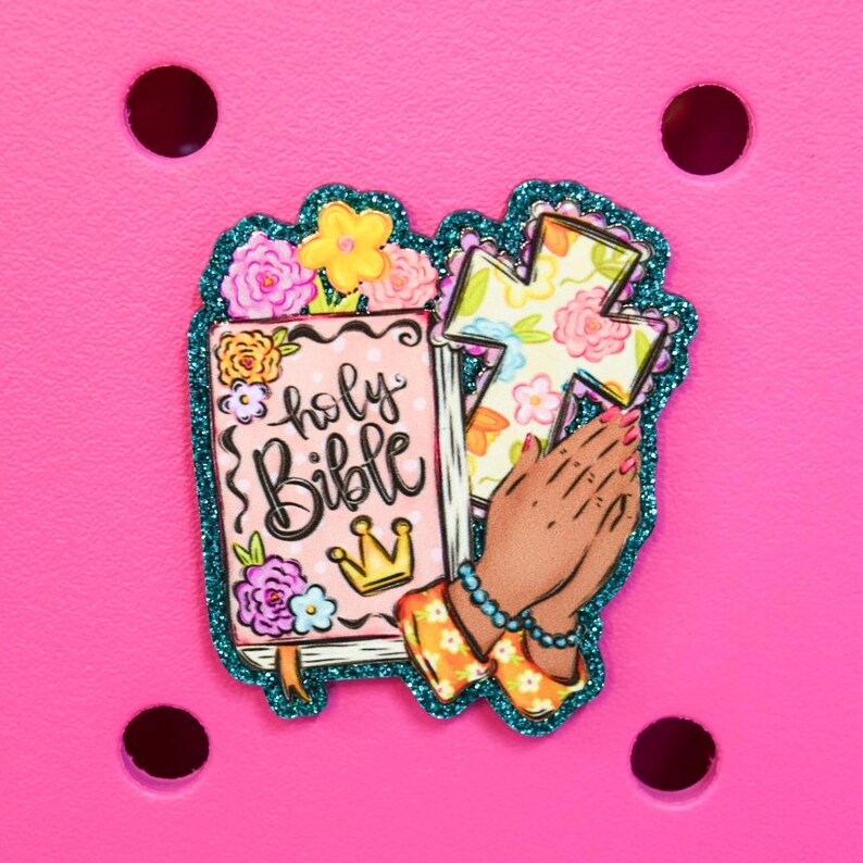 May include: A decorative charm featuring a pink Bible with the words "holy Bible" and a gold crown. The charm also includes a floral cross and praying hands, all outlined in teal glitter. The charm is set against a bright pink background.