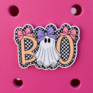 May include: A decorative shoe charm featuring a white ghost character centered between the letters B and O. The letters and ghost are outlined in a black and white checkerboard pattern. The ghost wears a purple bow, and the letters have pink bows.