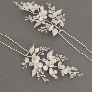 May include: Two white floral hair pins with delicate leaves, small flowers, and crystal accents. The pins are made of silver-colored wire and are arranged on a gray background. These hair accessories are suitable for weddings or special occasions.
