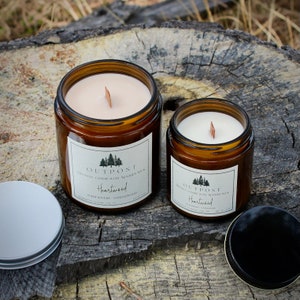 May include: Two amber glass jars with lids containing soy wax candles. The candles are labeled "Outpost" and "Heartwood" with a wooden wick. The larger jar is labeled "Wildflowers, Sandalwood".